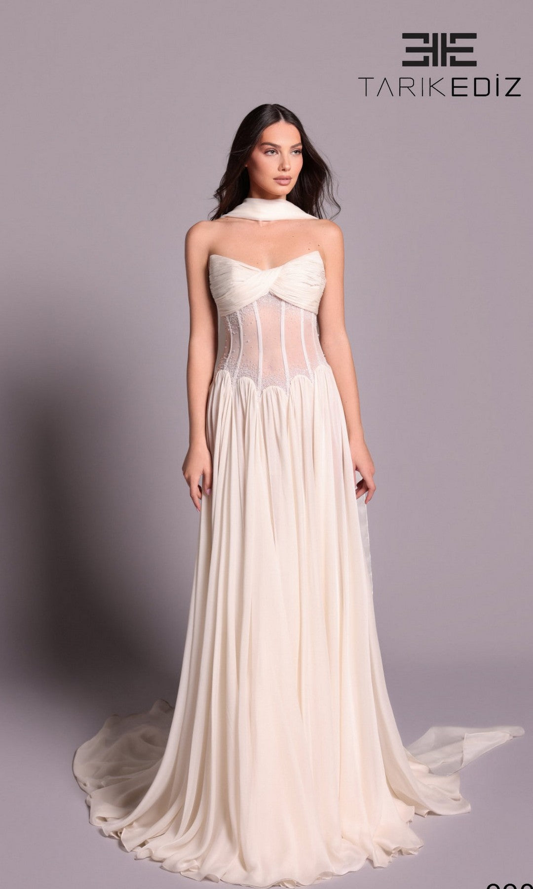 A woman in the Tarik Ediz 99076 long formal dress, featuring a strapless neckline, sheer corset bodice, and floor-length vanilla chiffon skirt, stands before a plain backdrop. The TARIK EDIZ logo is visible in the upper right corner.